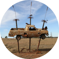 The image is of an art sculpture that shows a rusty ute being hoisted by three poles. The background sky is blue and ground is dry and brown. The image is of an art sculpture that shows a rusty ute being hoisted by three poles. The background sky is blue and ground is dry and brown.